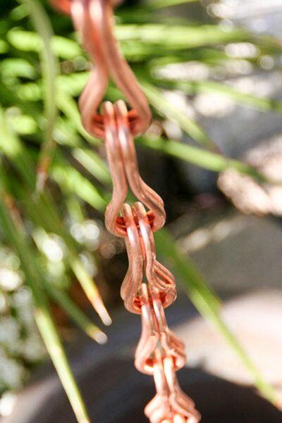 Stylish copper figure 8 rain chain with greenery in the background. Eight Shape Link Copper Rain Chain. Figure 8 design. 8ft section. 1-1/8" x 3-5/8" links. Pure copper. Living patina. No assembly. Contractor DIY. Damascus OR.