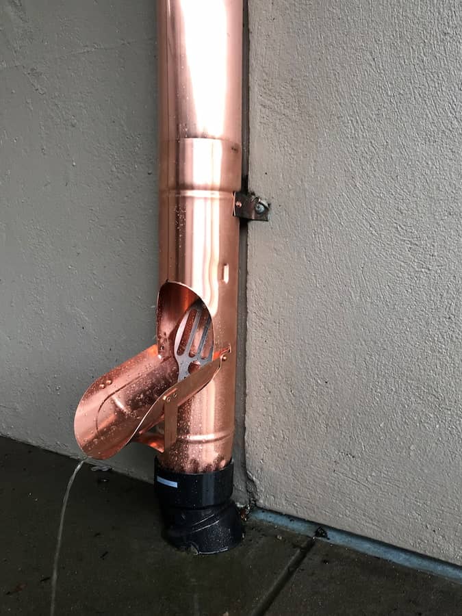 Shiny copper downspout attached to exterior wall, featuring a curved cleanout section fitted with a debris screen at the bottom over a concrete surface—demonstrating professional gutter maintenance and effective downspout filtration.
