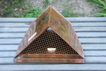 Triangular copper leaderhead with mesh screen displayed on wooden surface.