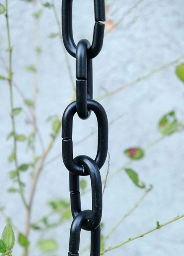 Black aluminum rain chain installed outdoors with greenery in the background.