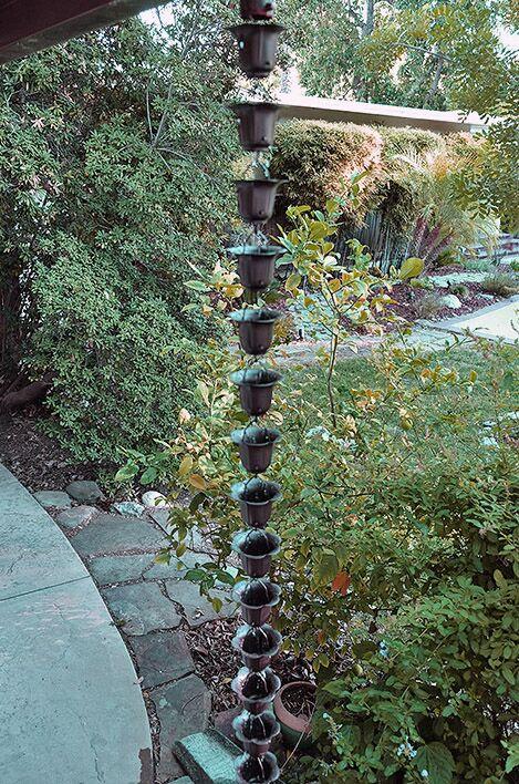 Long dark bronze buttercup rain chain hanging in a lush garden setting.