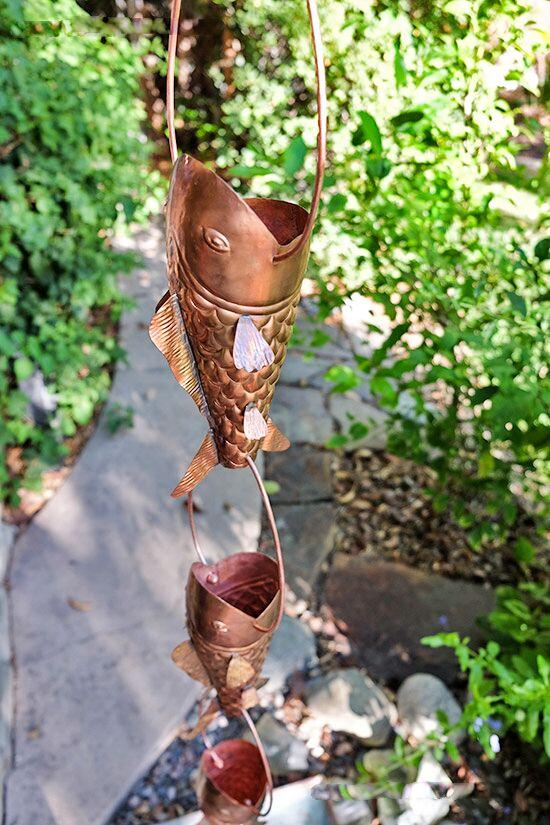 Copper fish rain chain hanging outdoors with lush greenery in the background. Koi Fish Copper Rain Chain. Sculptural swimming design. 8ft section. 8 fish. 9.5" length. 4" mouth. Detailed scales/fins. V-hook included. Contractor DIY. Damascus OR.
