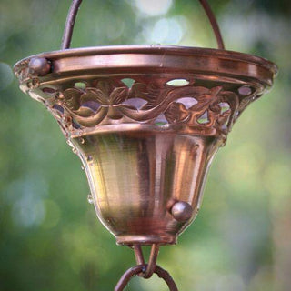 Copper floral bell cup rain chain with water droplets, set against a blurred outdoor background.