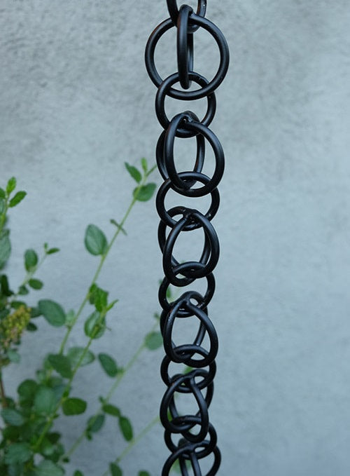 Black hooked rings rain chain displayed vertically with a gray wall and greenery.