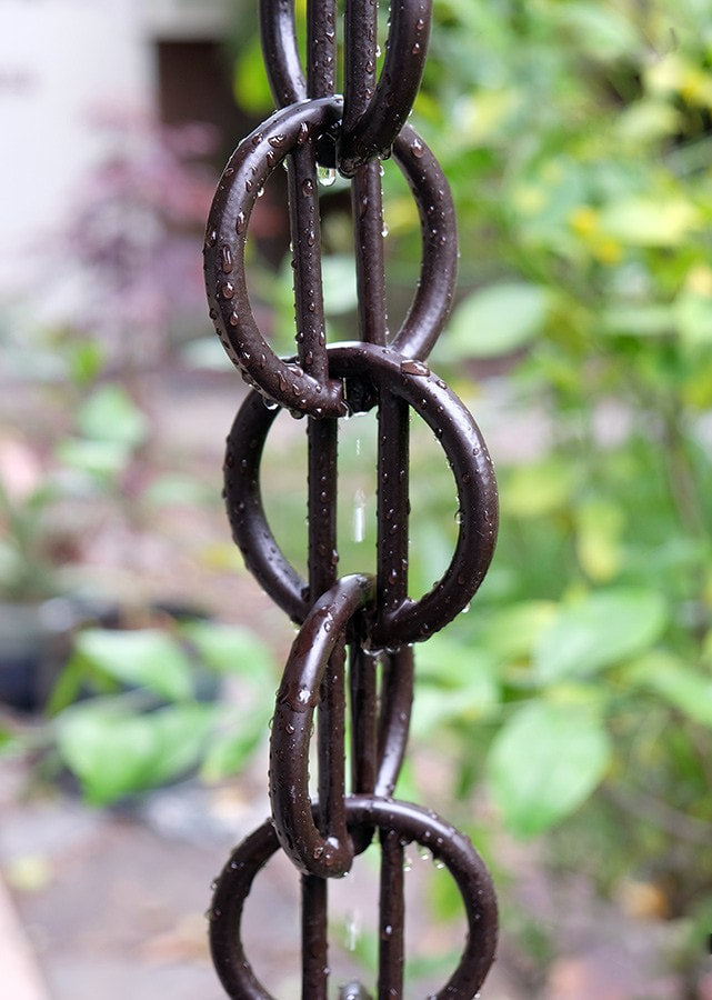 Black rain chain with water droplets, set against a garden backdrop.