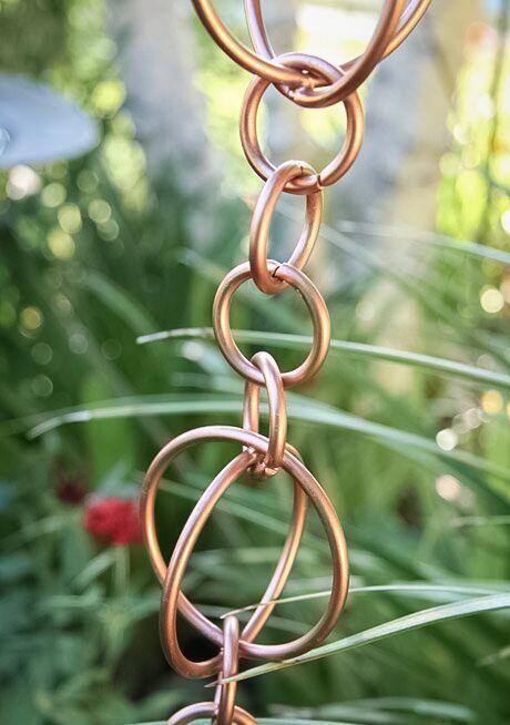 Copper hoops link rain chain in a garden setting with greenery.