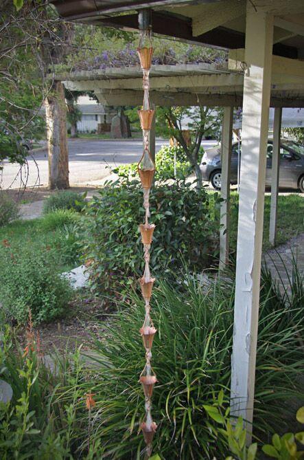 Full view of a tulip copper cup rain chain hanging from a roof in a lush garden.