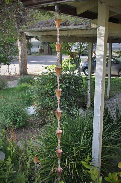 Full view of a tulip copper cup rain chain hanging from a roof in a lush garden.