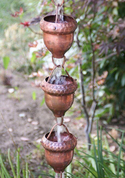 Decorative copper oaknut rain chain with water flowing in a garden setting.