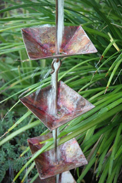 Multiple shallow square cups of a copper rain chain cascading water in a garden setting.