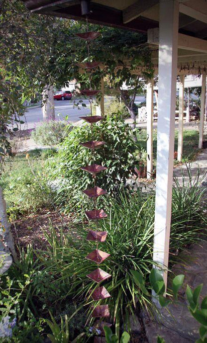 Full view of a shallow square cup rain chain installed on a porch, surrounded by greenery.