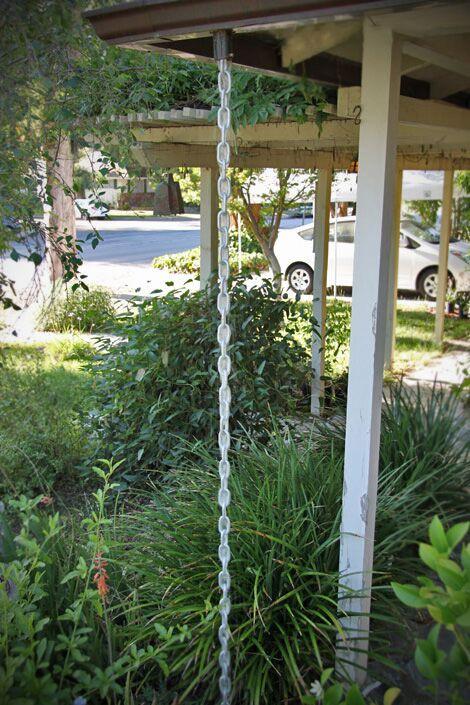 Full view of a silver rain chain installed on a house with a garden.