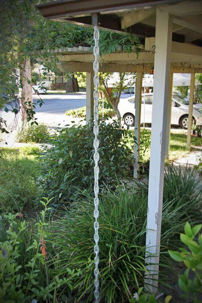 Full view of a silver rain chain installed on a house with a garden.