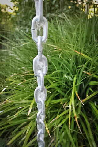 Silver rain chain in use, guiding water with greenery in the background.
