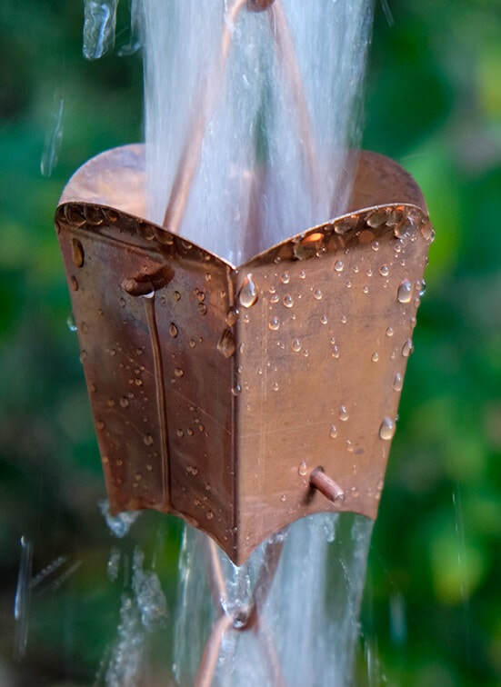 Square tulip cup rain chain in copper with water cascading, set against greenery.
