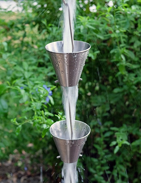Stainless steel rain chain with water cascading through cups in a garden setting.
