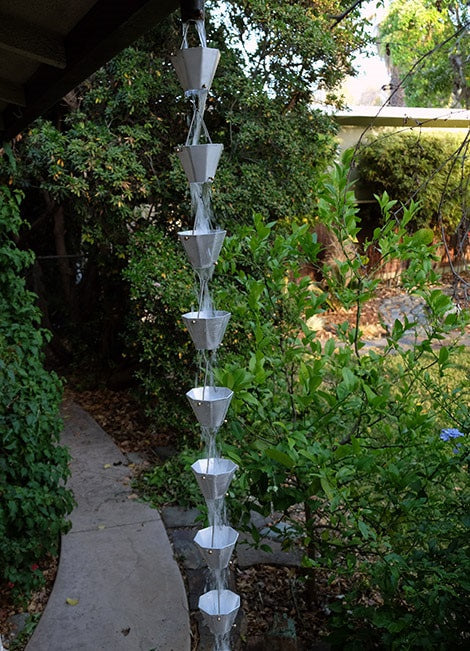 Full view of an octagonal aluminum rain chain with cascading water in a garden setting.